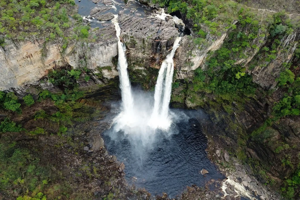 Foto de uma das cachoeiras da Chapada dos Veadeiros, com duas quedas d'água fortes e altas que desaguam num riacho. Há pedras ao redor e muita vegetação verde.