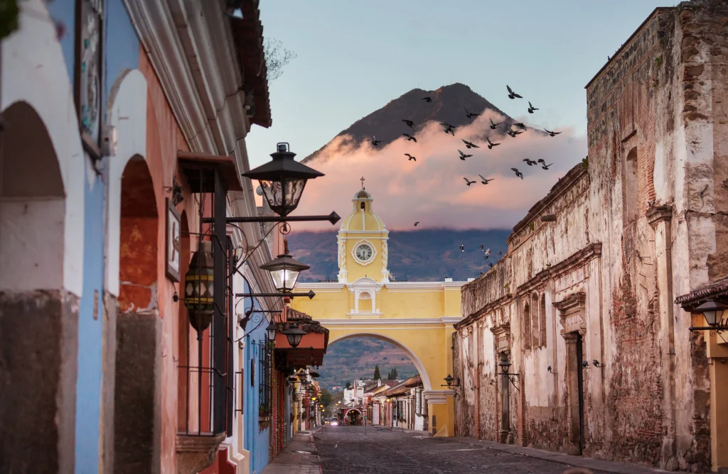 Foto da cidade de La Antigua, na Guatemala, com o morro ao fundo rodeado por nuvens. O céu está azul. Na parte da frente podemos ver as construções antigas da cidade, com um portal amarelo e ruas de paralelepípedos.