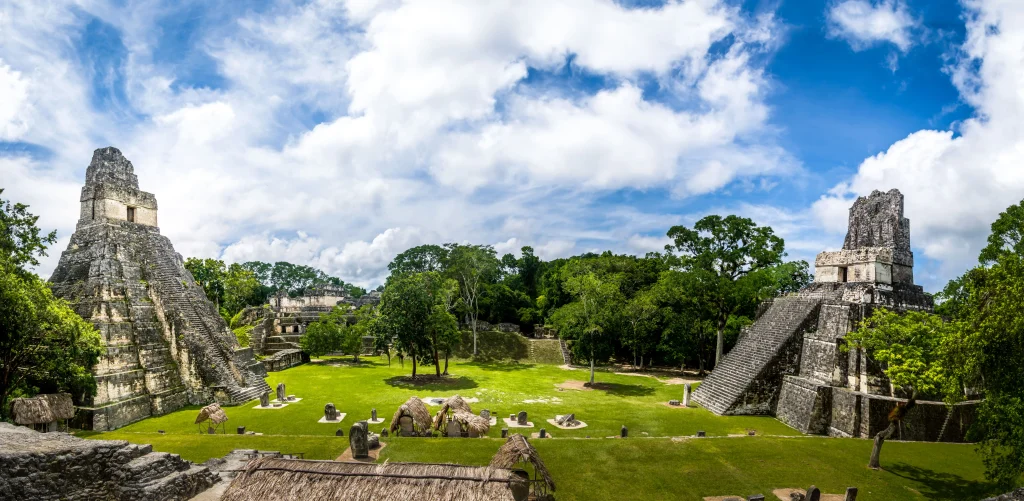 Foto do complexo onde ficam os monumentos parecidos com pirâmides de Tikal, que foi um dos maiores centros populacionais e culturais da civilização maia. A vegetação é bem verde e o céu azul. Não há pessoas na foto.
