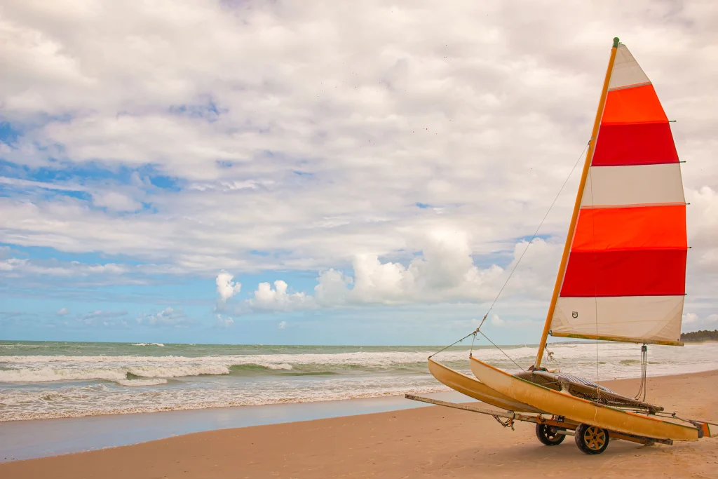 Foto de um barco estacionado na faixa de areia da uma das praias de Maceió, no Nordeste do Brasil. O céu está azul e o mar está calmo ao fundo. O barco possui uma vela com uma bandeira nas cores vermelho, laranja e branco.