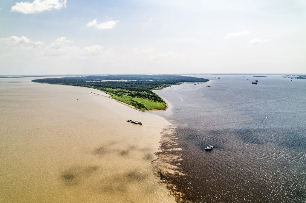 Foto incrível do encontro dos rios em Manaus, no Amazonas. É possível ver o contraste de cores do marrom e as águas se encontrando. Há alguns barcos na imagem e um território de terra e vegetação ao centro.