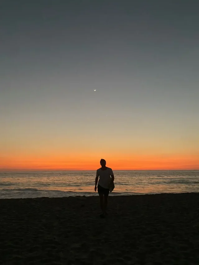 Foto com a silhueta de um homem contrastando com o pôr do sol na praia de Zicatela, onde as pessoas surfam.