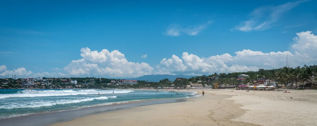 Águas claras e praia de areia branca, com céu azul e alguns surfistas, ilustrando as melhores coisas para fazer em Puerto Escondido.