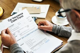 The image shows a close-up of an individual's hands holding a "Car Insurance Claim Form". The form is partially filled out and rests on a wooden table. The person is wearing glasses and has a silver watch on the left wrist.