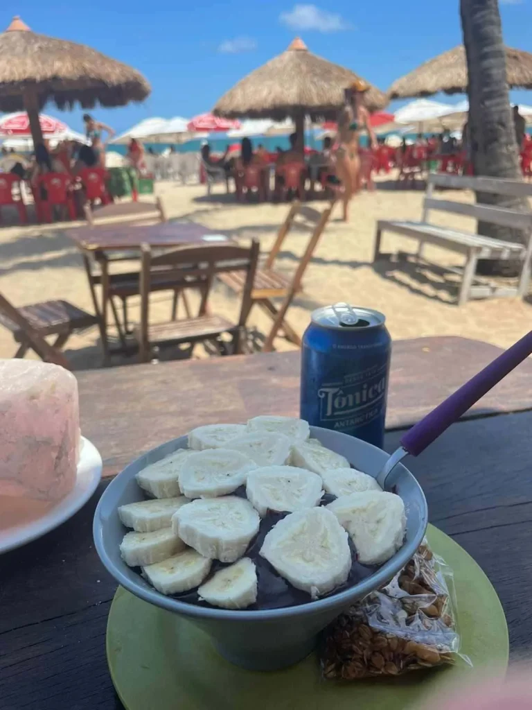 Foto de um prato típico do Brasil: o açaí. Há açaí e bananas cortadas por cima. Além disso, há amendoins ao lado do prato e uma lata de refrigerante. O céu está azul.