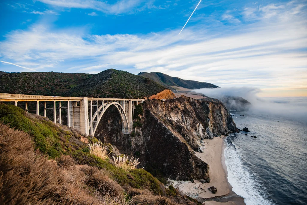 Foto de uma ponte da Big Sur, na Califórnia, Estados Unidos. Ao fundo tem nuvens acima do mar, o mar com ondas, uma faixa de areia, o céu bem azul e a vegetação cobrindo toda a montanha.