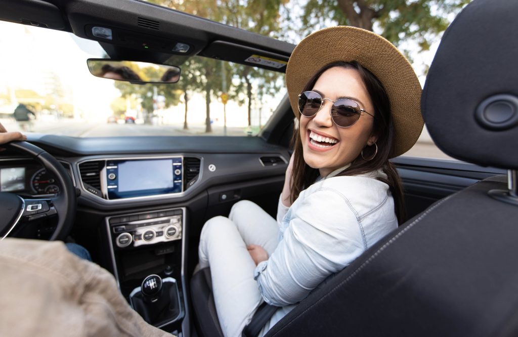Mulher sorrindo para câmera com chapéu na cabeça e óculos de sol. Ela está sentada no banco passageiro de um carro conversível.