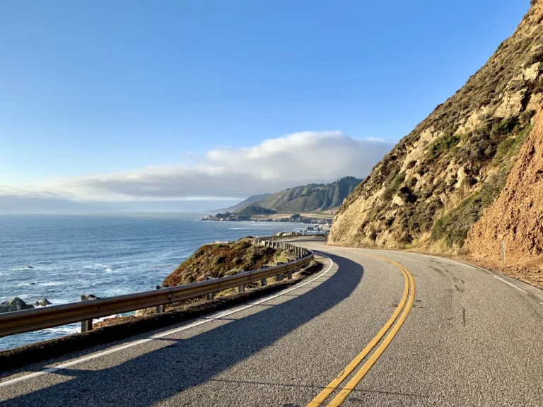 Foto de uma ponte da Big Sur, na Califórnia, Estados Unidos. Ao fundo tem nuvens acima do mar, o mar com ondas, uma faixa de areia, o céu bem azul e a vegetação cobrindo toda a montanha.