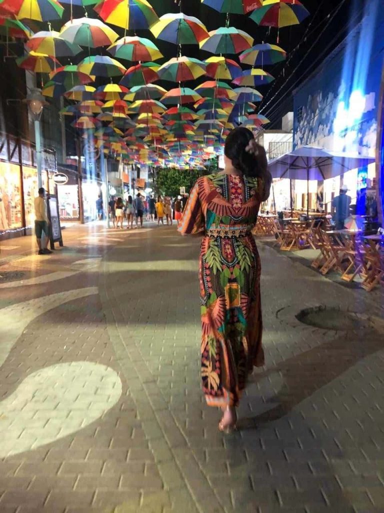 Foto de uma mulher andando pelo centro da vila de Porto de Galinhas. Há vários guarda-chuvas coloridos que fazem uma espécie de teto durante toda a rua. Há pessoas nos restaurantes nas laterais da rua.