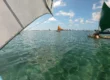 Foto do mar bem verde cristalino com as jangadas sobre ele. Há pessoas nas jangadas e as jangadas são coloridas. O céu está bem azul.