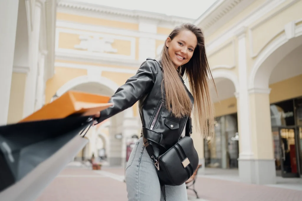 Foto de uma mulher sorridente olhando para câmera. Ela está segurando sacolas de compras e está dentro de um estabelecimento estilo shopping.