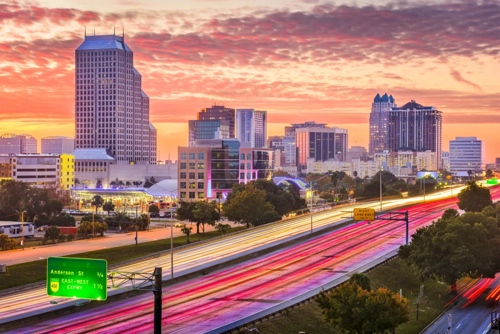 Foto de um lindo entardecer na cidade de Orlando, Flórida, Estados Unidos. Os prédios cinzas entram em contraste com o céu cor-de-rosa. As ruas registram as luzes dos carros que passam em alta velocidade.