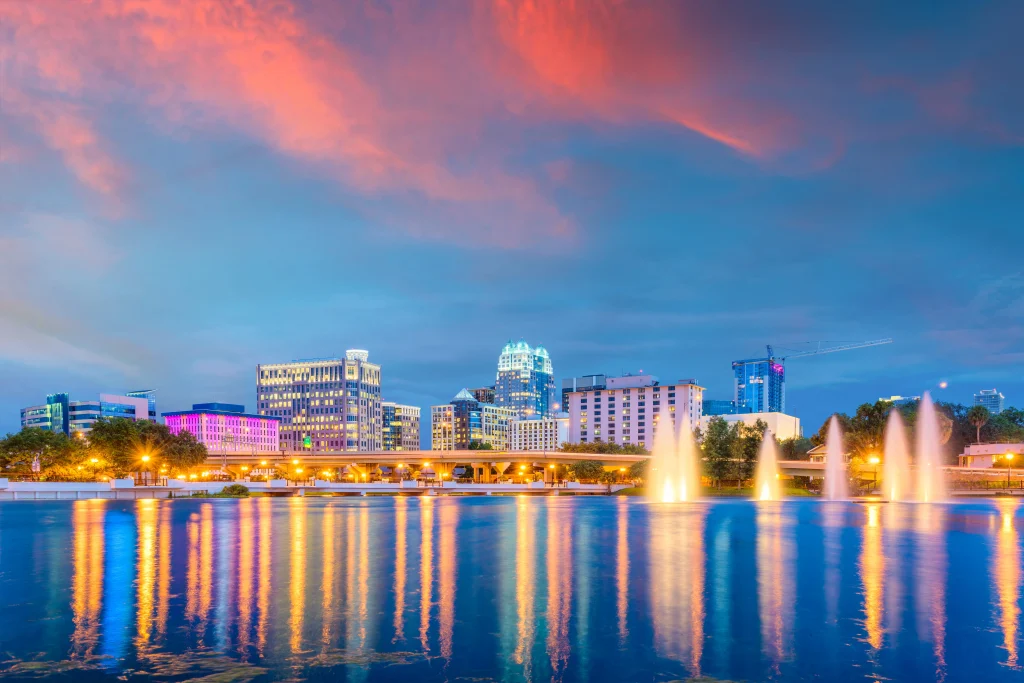 Foto de um lindo entardecer na cidade de Orlando, Flórida, Estados Unidos. Os prédios cinzas entram em contraste com o céu cor-de-rosa e azul. As luzes da cidade refletem no lago em frente aos prédios.