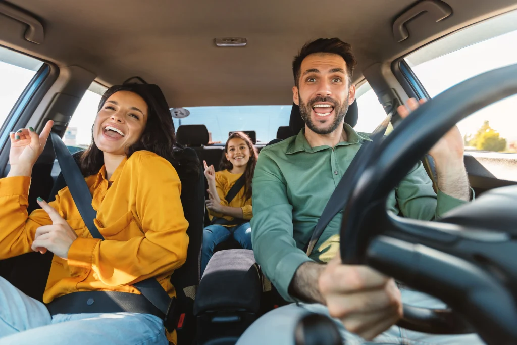 Uma mulher com camisa amarela e um homem com camisa verde estão felizes e sorrindo dentro de um carro durante o dia. No banco do passageiro está uma menina de blusa amarela que também sorri.