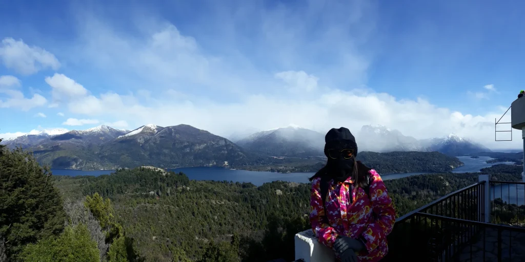 Uma mulher com gorro, óculos de sol e cachecol e ao fundo uma paisagem com céu azul, nuvens, montanhas e muitas árvores.