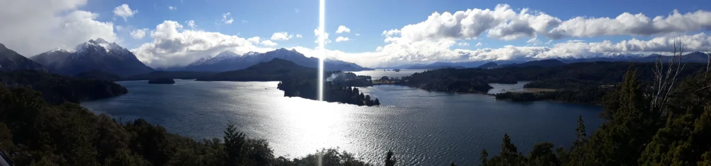 Uma imagem no formato horizontal de uma paisagem em Bariloche com lagos, árvores e montanhas.