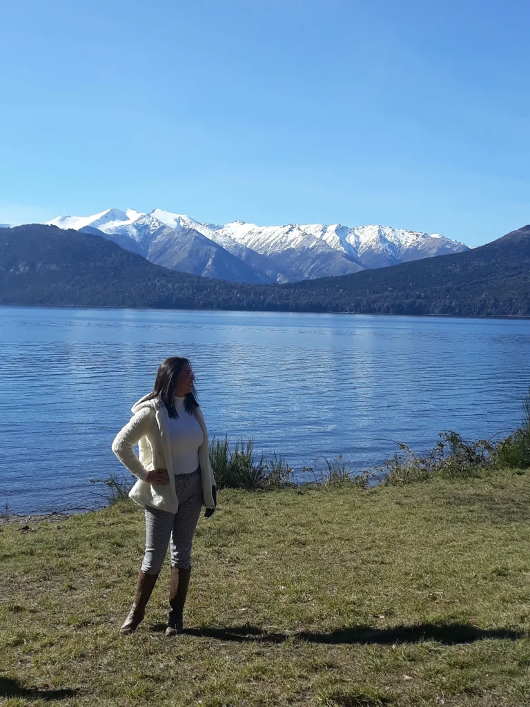 Uma mulher virada de lado com roupas em tons claros de pé e ao fundo uma paisagem com um lago e montanhas com gelo.