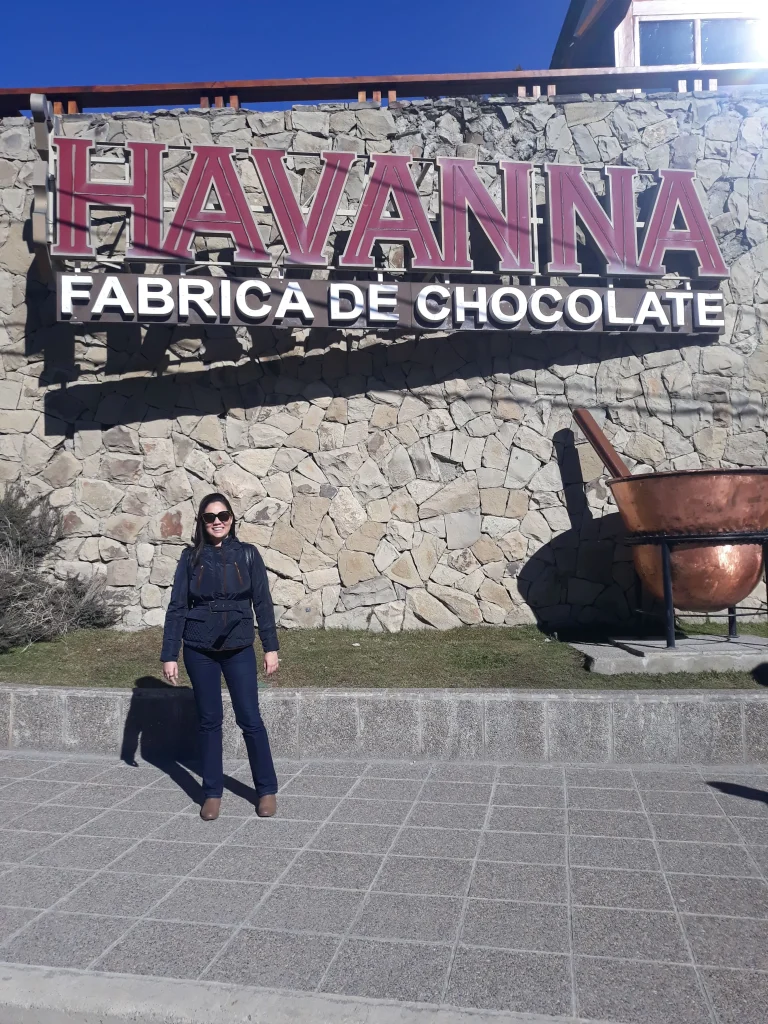 Uma mulher de roupa escura e óculos de sol ao centro da imagem e a frente de uma fachada de uma fábrica de chocolate em Bariloche.