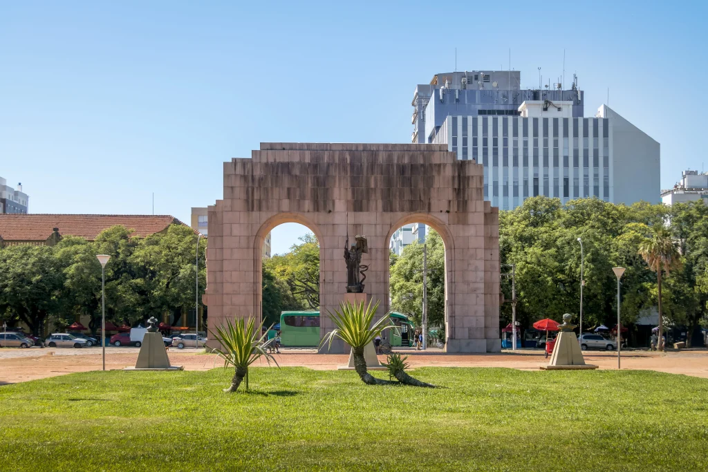 Uma imagem do monumento da Farroupilha no Parque da Redenção em Porto Alegre, Rio Grande do Sul. O céu está azul e há prédios atrás do monumento.