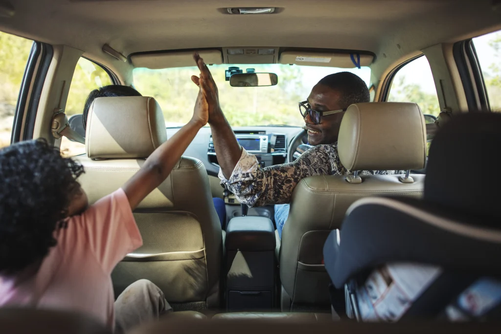 Um carro interno com uma família. Um homem está virado com a mãe estendida para cumprimentar de forma amigável a mão de uma menina que está sentada no banco do passageiro.