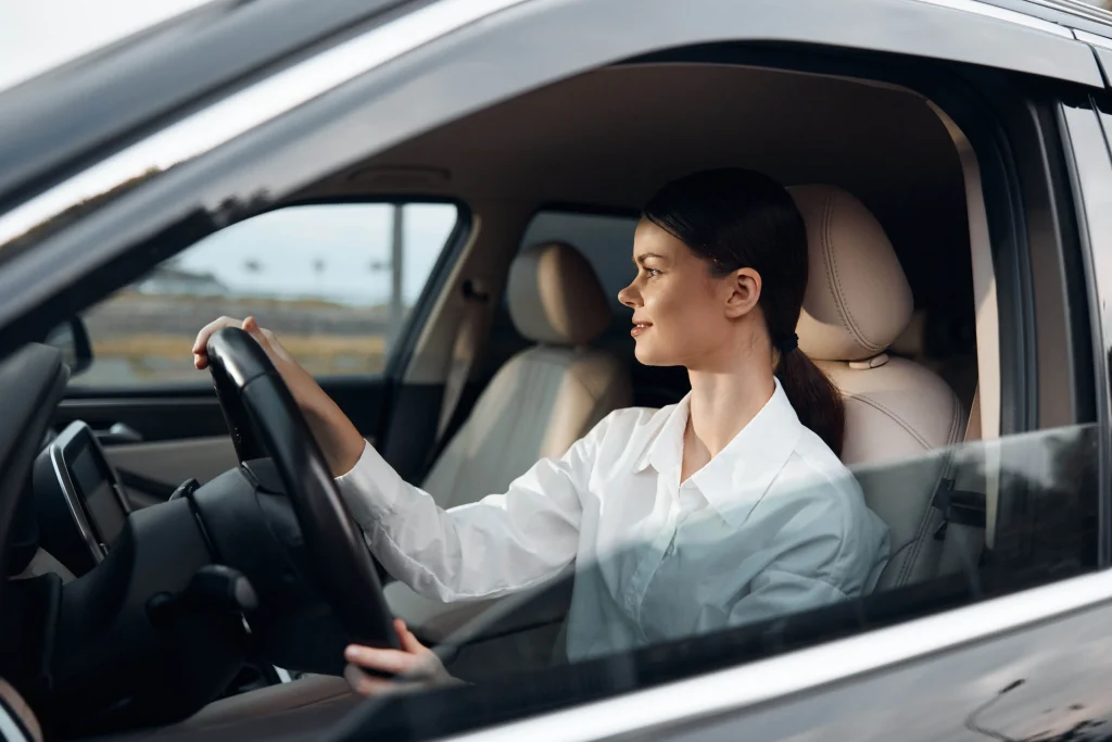 Uma mulher de cabelo preto preso e camisa branca está dentro do carro dirigindo e olhando para o horizonte