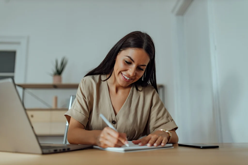 Uma mulher de cabelos escuros está sentada em uma mesa sorrindo enquanto escreve no seu caderno e ao seu lado está um computador.