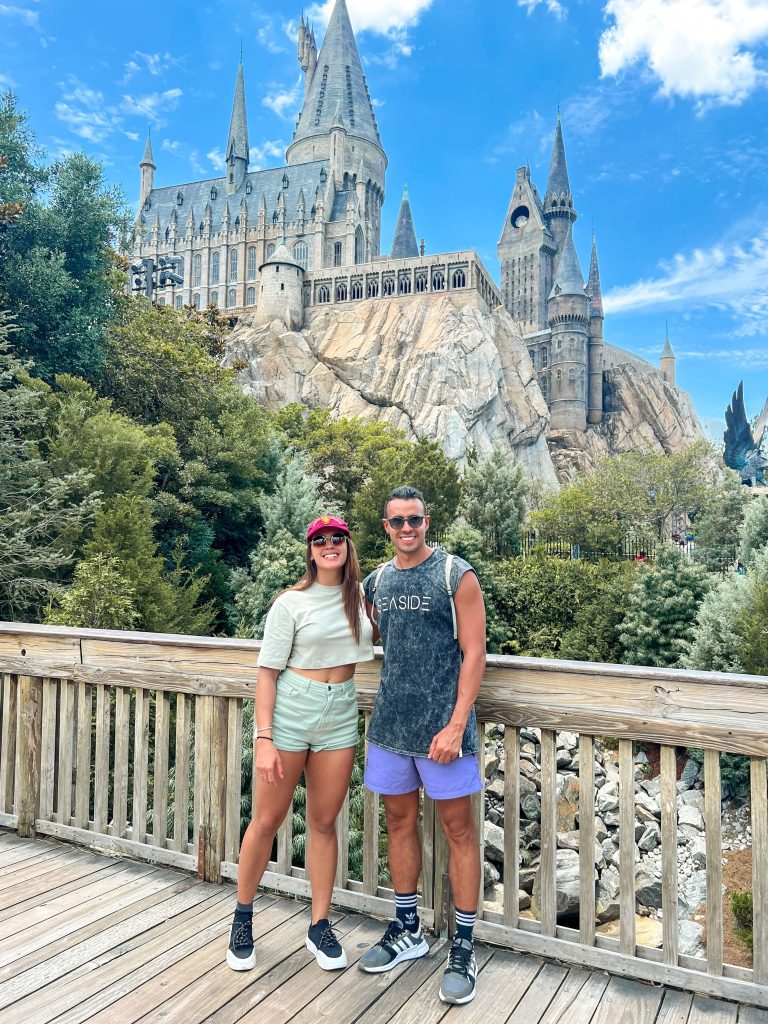 A foto mostra um casal sorridente para foto. Estão abraçados. Atrás deles tem a réplica de um dos castelos mais famosos dos filmes Harry Potter presente nos parques temáticos de Orlando. O céu está azul.