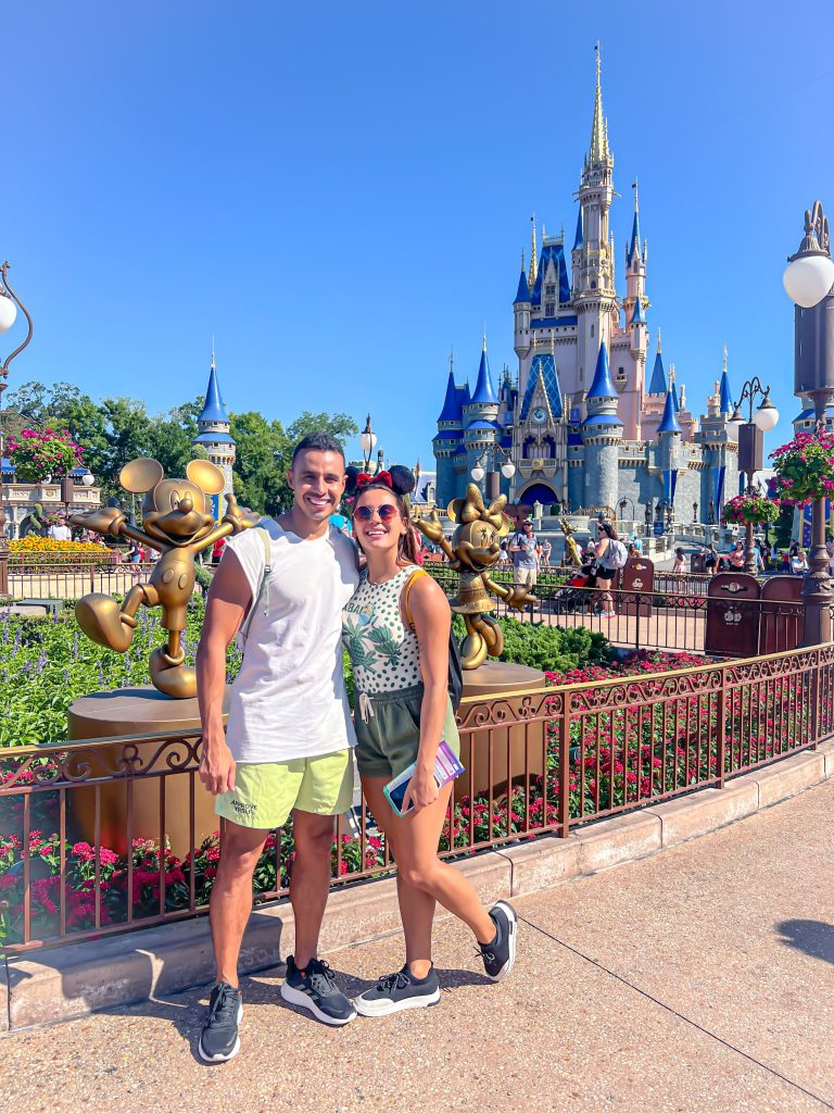 A foto mostra um casal sorridente para foto. Estão abraçados. Atrás deles tem o famoso castelo da Disney presente nos parques temáticos de Orlando. O céu está azul.