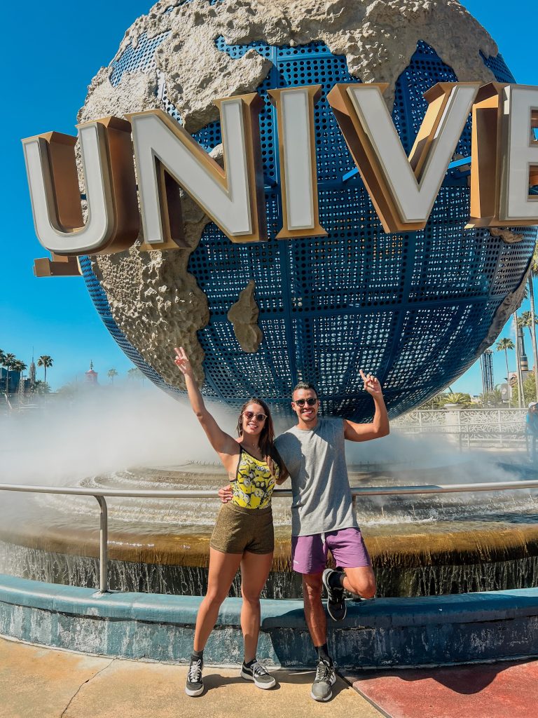 A foto mostra um casal sorridente para foto, estão abraçados e fazendo o número dois com a mão. Atrás deles tem o globo enorme escrito Universal presente nos parques temáticos de Orlando. O céu está azul.