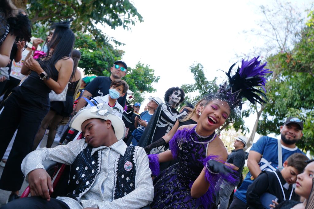 Joselito Carnaval 'dormido' en medio de la fiesta del Carnaval de Barranquilla, mientras una bailarina vestida de morado y negro celebra con alegría. Al fondo, la multitud refuerza el ambiente vibrante del evento.