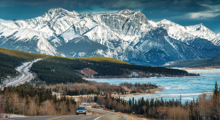Carro vindo em uma estrada das montanhas rochosas do Canadá com neve no topo e um lago do lado