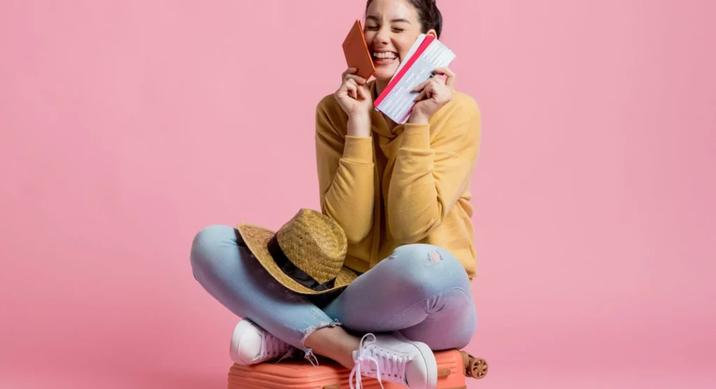 Mulher sorridente sentada em fundo rosa, segurando um cartão de crédito na mão e um celular, sugerindo compra online ou pagamento seguro.