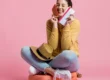 Mulher sorridente sentada em fundo rosa, segurando um cartão de crédito na mão e um celular, sugerindo compra online ou pagamento seguro.