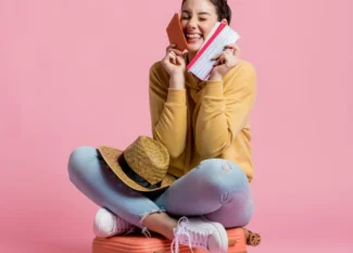Mulher sorridente sentada em fundo rosa, segurando um cartão de crédito na mão e um celular, sugerindo compra online ou pagamento seguro.