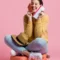 Mulher sorridente sentada em fundo rosa, segurando um cartão de crédito na mão e um celular, sugerindo compra online ou pagamento seguro.