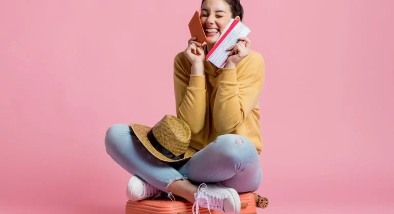 Mulher sorridente sentada em fundo rosa, segurando um cartão de crédito na mão e um celular, sugerindo compra online ou pagamento seguro.