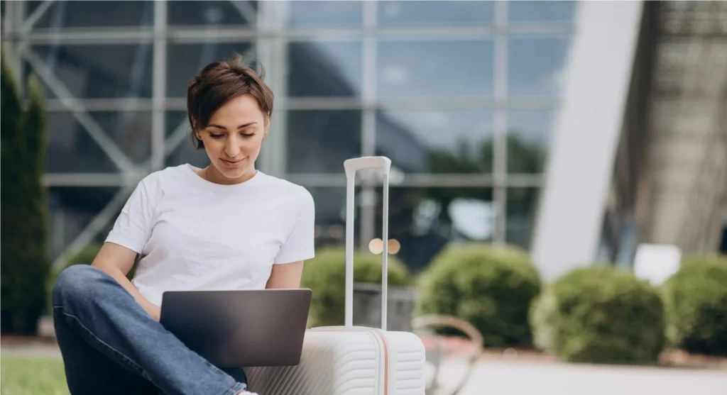 mulher sentada com um laptop no colo e uma mala de viagem ao lado