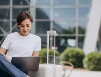 mulher sentada com um laptop no colo e uma mala de viagem ao lado