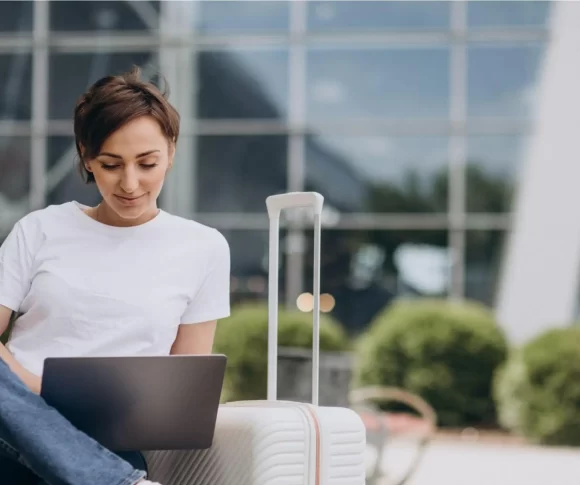 mulher sentada com um laptop no colo e uma mala de viagem ao lado