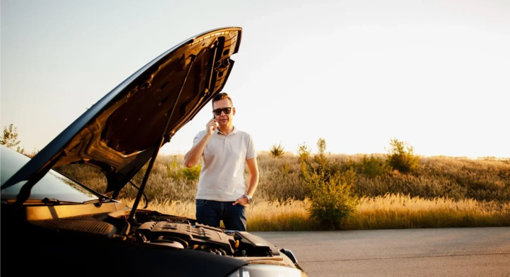 Homem em pé ao lado de um carro com o capô aberto, levantando a mão em gesto de conversa enquanto o motor fica exposto, em uma estrada ao ar livre ao pôr do sol.