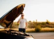 Homem em pé ao lado de um carro com o capô aberto, levantando a mão em gesto de conversa enquanto o motor fica exposto, em uma estrada ao ar livre ao pôr do sol.