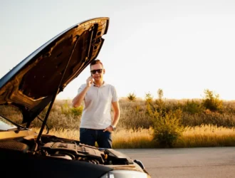 Homem em pé ao lado de um carro com o capô aberto, levantando a mão em gesto de conversa enquanto o motor fica exposto, em uma estrada ao ar livre ao pôr do sol.