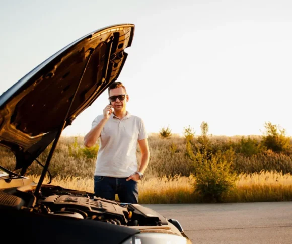 Homem em pé ao lado de um carro com o capô aberto, levantando a mão em gesto de conversa enquanto o motor fica exposto, em uma estrada ao ar livre ao pôr do sol.