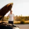 Homem em pé ao lado de um carro com o capô aberto, levantando a mão em gesto de conversa enquanto o motor fica exposto, em uma estrada ao ar livre ao pôr do sol.