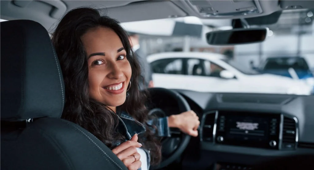 A smiling woman at the wheel of a modern car, with the dashboard and center console in the background, in an urban setting or a parking lot with other vehicles.