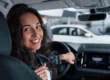 A smiling woman at the wheel of a modern car, with the dashboard and center console in the background, in an urban setting or a parking lot with other vehicles.