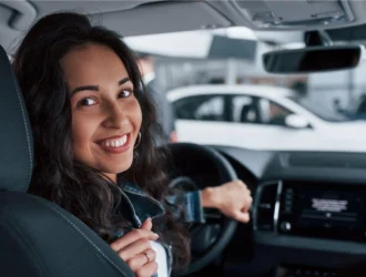 A smiling woman at the wheel of a modern car, with the dashboard and center console in the background, in an urban setting or a parking lot with other vehicles.