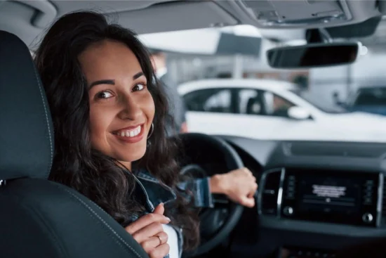 A smiling woman at the wheel of a modern car, with the dashboard and center console in the background, in an urban setting or a parking lot with other vehicles.