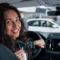 A smiling woman at the wheel of a modern car, with the dashboard and center console in the background, in an urban setting or a parking lot with other vehicles.