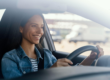 Mulher sorrindo ao dirigir um carro, com o volante nas mãos e expressão de tranquilidade, ao lado do banco do motorista, em ambiente externo com paisagem desfocada ao fundo.
